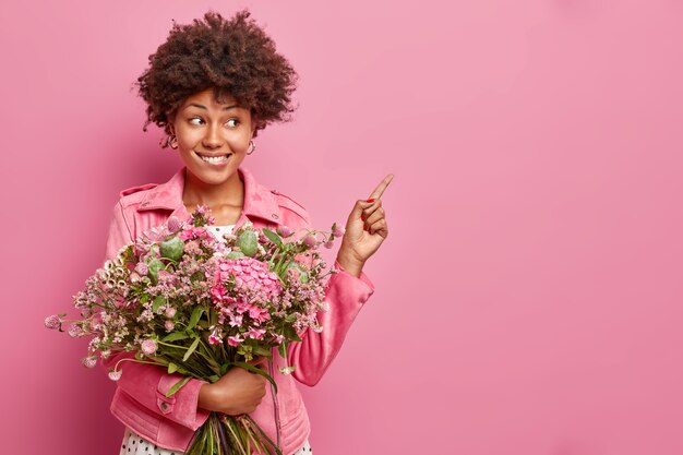 Positif jeune femme frisée pose avec joli bouquet de fleurs points à l'espace vide montre le contenu publicitaire porte veste isolé sur mur rose
