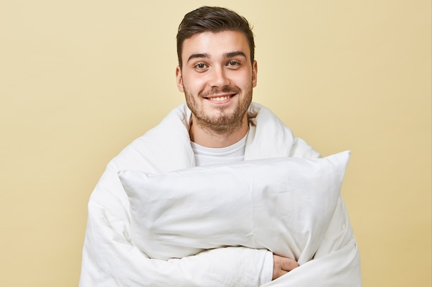 Positif jeune homme gai avec un sourire mignon et un visage mal rasé debout au mur blanc, enveloppé dans une couverture blanche, se sentant ravi, se remettant du froid, tenant un oreiller, s'endormir dans son lit