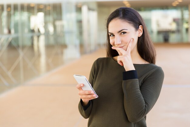 Positive femme latine timide avec téléphone portable touchant le visage