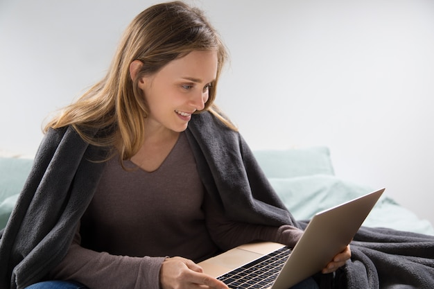 Positive jeune femme à l&#39;aide d&#39;un ordinateur portable dans la chambre