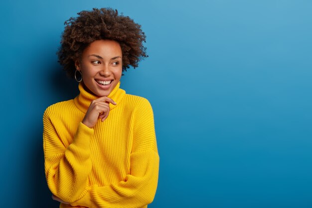 Positive jeune femme bouclée vêtue d'un pull confortable jaune, tient le menton, regarde de côté avec une expression rêveuse, a une idée intéressante à l'esprit, isolée sur fond bleu.