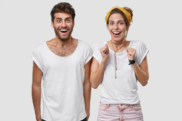 Positive jeune femme de race blanche serre les poings, porte un t-shirt, regarde avec impatience, se tient près de l'homme joyeux, célèbre le succès et le triomphe