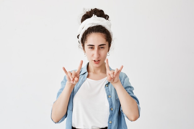 Positive jeune fille sombre avec des cheveux foncés et ondulés en chignon, portant du chiffon s'amusant, des lèvres boudeuses, montrant un signe de rock avec les deux mains. Concept de langage corporel et de gestes.