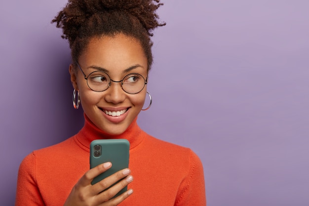 Positive jolie jeune femme afro-américaine concentrée de côté, porte des lunettes rondes et un pull décontracté, sourit joyeusement