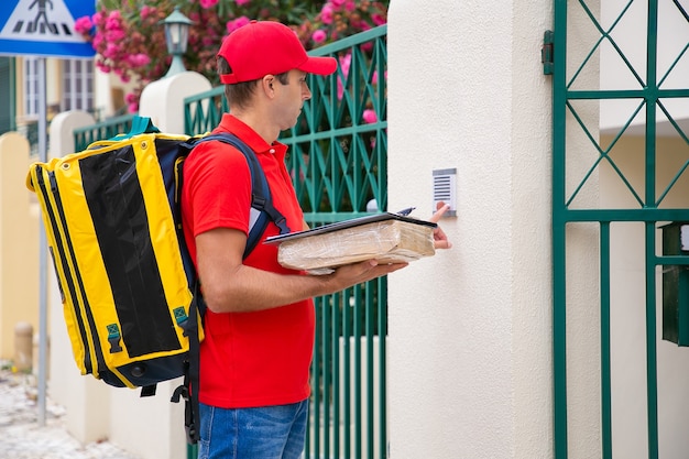 Photo gratuite postier concentré sonnant à la porte et tenant le colis et le presse-papiers. beau courrier en uniforme rouge portant un sac à dos, debout à l'extérieur et livrant la commande. service de livraison et concept de poste