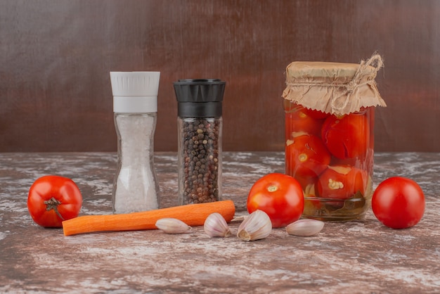 Un pot de tomates marinées, de grains de poivre et de légumes frais sur table en marbre.