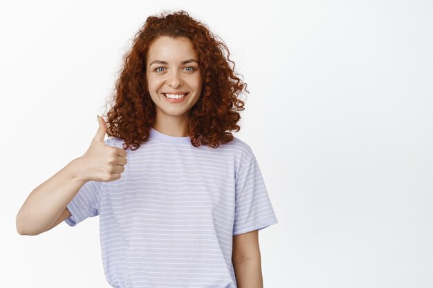 Pouce en l'air fille. Femme souriante et souriante aux cheveux bouclés montrant comme, geste d'approbation, vous félicitant, excellent choix, recommandant qch sur blanc.