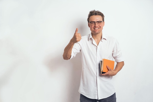 Pouce en l'air homme indépendant souriant dans une chemise blanche tenant un cahier ou un journal orange isolé sur fond blanc ou au bureau Portrait d'un jeune homme d'affaires positif tenant son journal