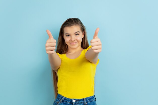 Pouces vers le haut. Portrait de l'adolescente caucasienne isolée sur le mur bleu. Beau modèle en tenue jaune décontractée. Concept d'émotions humaines, expression faciale, ventes, publicité. Copyspace. Ça a l'air mignon.