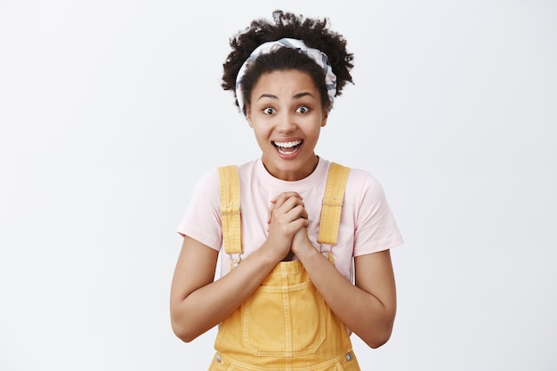 Pour de vrai, est-ce pour moi. Portrait de touché surpris et heureux heureux femme afro-américaine en salopette élégante jaune applaudissant des paumes sur la poitrine et souriant largement avec un regard reconnaissant