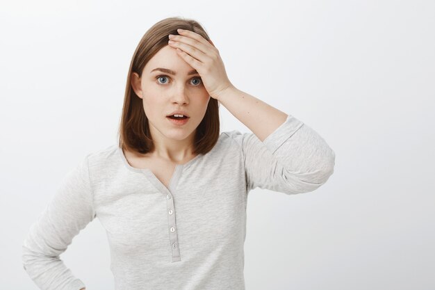 Pourquoi moi mon dieu. Portrait de jeune étudiante embarrassée et inquiète debout dérangée et inquiète sur un mur gris en chemisier blanc décontracté tenant la paume sur le front se plaignant de la vie difficile