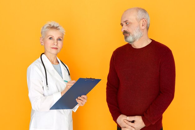 Praticien de femme mûre sérieuse réussie en blouse blanche vérifiant l'état de santé avec son patient de sexe masculin âgé au cours de l'examen physique, tenant le presse-papiers et le stylo, regardant la caméra