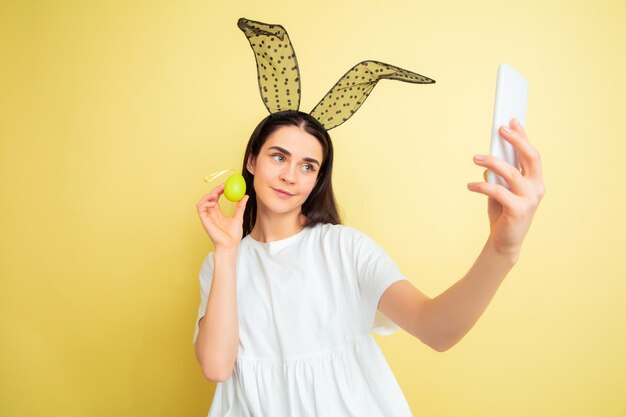 Prendre selfie. Femme de race blanche comme un lapin de Pâques sur fond de studio jaune. Bonnes salutations de Pâques. Beau modèle féminin. Concept d'émotions humaines, expression faciale, vacances. Copyspace.