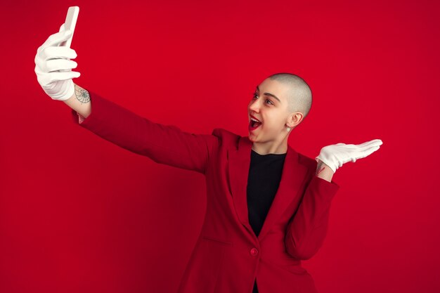 Prendre un selfie ou un vlog. Portrait de jeune femme chauve caucasienne isolée sur mur rouge. Beau modèle féminin en veste. Émotions humaines, expression faciale, ventes, concept publicitaire. Culture bizarre.