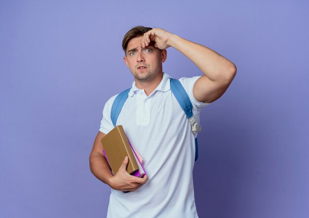 Préoccupé jeune beau étudiant masculin portant un sac à dos tenant des livres et mettant la main sur le front isolé sur un mur bleu