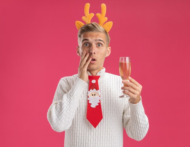 Préoccupé jeune beau mec portant bandeau de bois de renne et cravate de père Noël tenant un verre de champagne touchant le visage isolé sur un mur rose avec copie