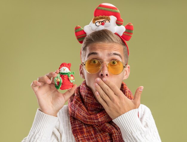 Préoccupé jeune beau mec portant bandeau et écharpe du père Noël regardant la caméra tenant l'ornement de Noël de bonhomme de neige en gardant la main sur la bouche isolé sur fond vert olive