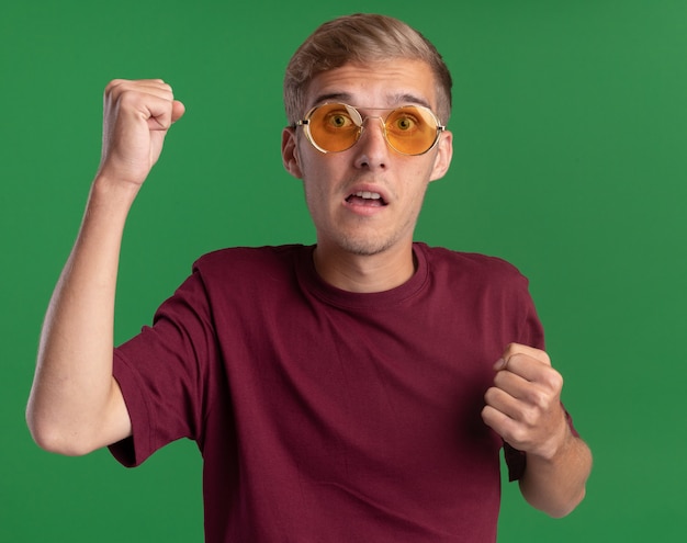 Préoccupé jeune beau mec portant chemise rouge et lunettes montrant oui geste isolé sur mur vert