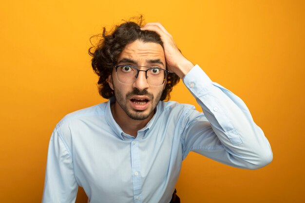 Préoccupé jeune bel homme caucasien portant des lunettes mettant la main sur la tête en regardant la caméra isolée sur fond orange