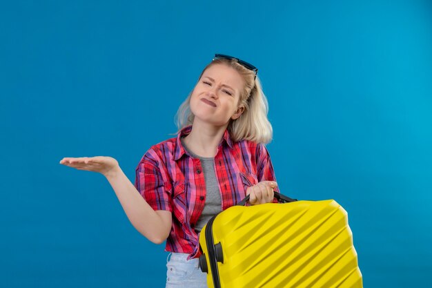 Préoccupé Jeune Femme Voyageur Portant Chemise Rouge Et Lunettes Sur La Tête Tenant Des Points De Valise à Côté Sur Un Mur Bleu Isolé