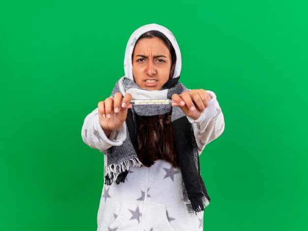 Photo gratuite préoccupé jeune fille malade mettant sur l'écharpe d'usure de capuche tenant le thermomètre à la caméra isolée sur fond vert