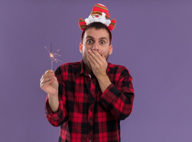 Préoccupé jeune homme de race blanche portant bandeau du père noël tenant un cierge magique en gardant la main sur la bouche isolé sur mur violet avec espace copie