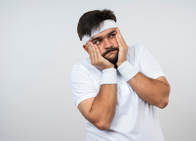 Préoccupé jeune homme sportif regardant côté portant bandeau et bracelet mettant les mains sur les joues isolé sur mur blanc