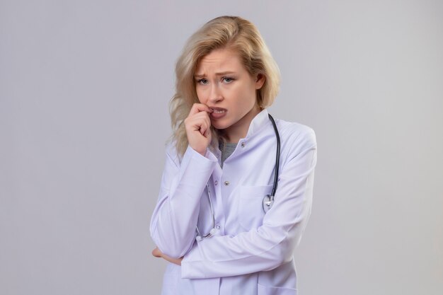 Préoccupé jeune médecin portant un stéthoscope en robe médicale mis la main sur la mâchoire sur le mur blanc