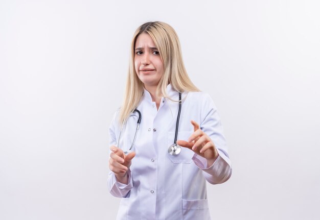 Préoccupé médecin jeune fille blonde portant un stéthoscope et une robe médicale montrant la taille avec les deux mains sur un mur blanc isolé