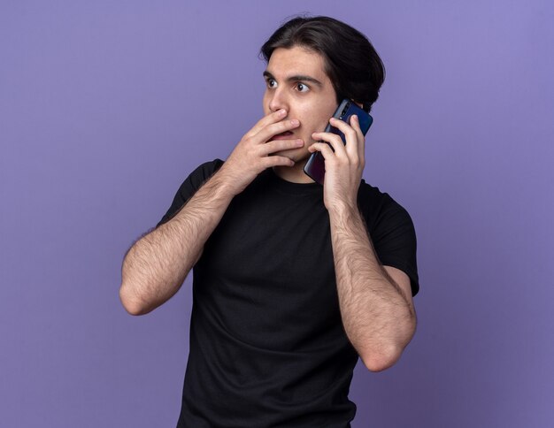 Préoccupé à la recherche de côté jeune beau mec portant un t-shirt noir parle au téléphone bouche couverte avec main isolé sur mur violet