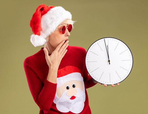 Préoccupée jeune femme blonde portant chapeau de Noël et pull de Noël du père Noël avec des lunettes en gardant la main sur la bouche tenant et regardant réveil isolé sur fond vert olive