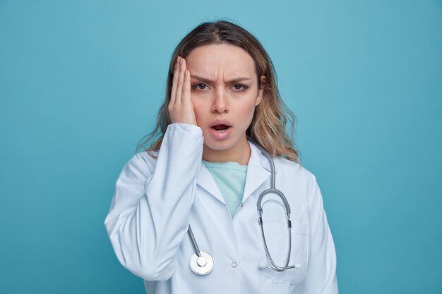 Préoccupée jeune femme médecin portant une robe médicale et un stéthoscope autour du cou en gardant la main sur le visage
