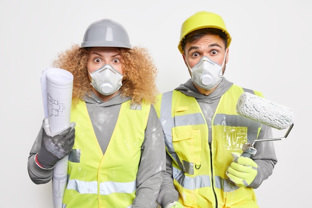 Prestation industrielle. Des femmes et des hommes choqués portant un masque de protection en uniforme de sécurité tiennent des outils de construction et un plan