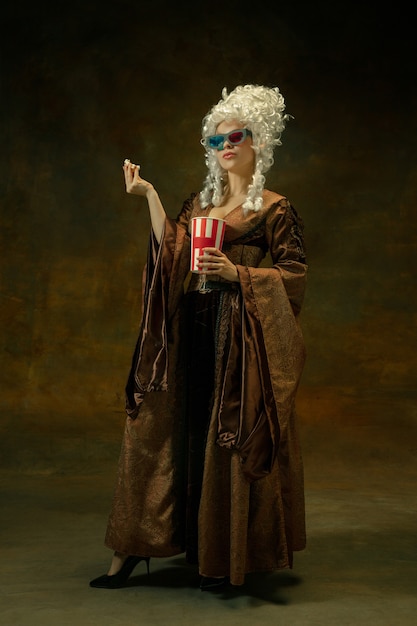 Prêt pour le cinéma. Portrait de femme médiévale en vêtements vintage avec lunettes 3D, pop-corn sur fond sombre. Modèle féminin en tant que duchesse, personne royale. Concept de comparaison des époques, de la mode, de la beauté.