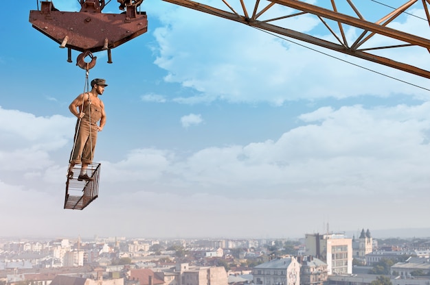 Prêt pour la journée. Portrait en contre-plongée d'un jeune constructeur rétro musclé posant sur une barre transversale suspendue à une grue sur un gratte-ciel