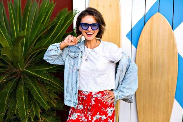 Printemps été Lumineux Positif Portrait De Femme Brune Souriante Heureuse Portant Une Tenue Hipster Féminine à La Mode Souriant Et S'amusant, Posant Devant Des Planches De Surf Et Des Palmiers.