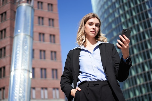 Photo gratuite prise de vue en angle bas d'une femme d'affaires en costume debout dans la rue et regardant un téléphone portable tenant intelligent