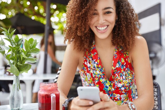 Prise de vue en extérieur d'une belle femme frisée à la peau foncée, lit des nouvelles agréables ou recherche des informations sur les réseaux sociaux