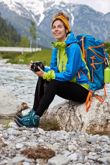 Prise de vue en extérieur d'une femme heureuse se repose alors qu'elle est assise sur une pierre près d'une petite rivière de montagne, tient un appareil photo professionnel pour prendre des photos