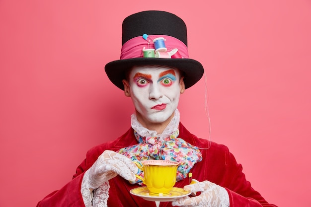 Prise de vue horizontale d'un chapeau masculin sérieux pose avec une tasse de thé porte un chapeau a des manières de robes de gentleman aristocratique pour les poses de carnaval de mascarade à l'intérieur