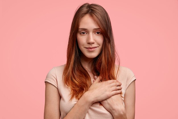 Prise de vue horizontale d'une femme à la peau amicale avec des taches de rousseur, garde les mains sur le cœur, regarde avec bonheur la caméra