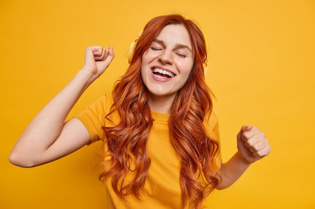 Prise de vue en intérieur d'une adolescente rousse heureuse se sentant détendue, secoue les bras au rythme de sa musique préférée porte des écouteurs sans fil profite d'un bon son sourit largement