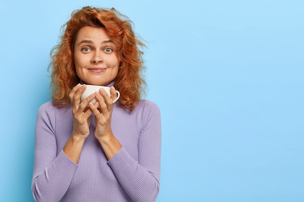 Prise de vue à l'intérieur d'une belle rousse a une pause-café, tient une tasse blanche avec une boisson aromatique, sourit et regarde, profite d'une belle conversation tout en buvant du thé le matin, discute des nouvelles