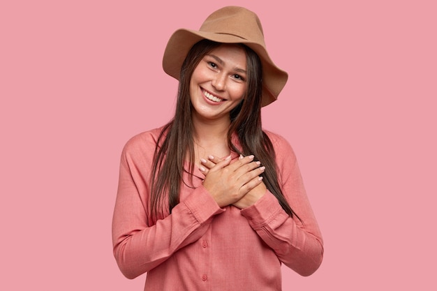 Prise de vue en intérieur d'une femme heureuse et touchée à l'air agréable a une apparence spécifique, garde les deux paumes sur le cœur