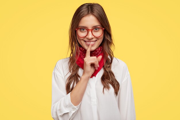 Prise de vue en intérieur d'une femme joyeuse satisfaite a les cheveux légèrement peignés, garde l'index sur la bouche