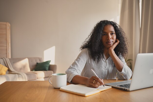 Prise de vue à l'intérieur d'une femme sérieuse belle jeune métisse indépendante aux cheveux ondulés travaillant à distance à l'aide d'un ordinateur portable, assise au bureau à domicile avec une tasse et un journal, écrit, fait des plans pour la journée