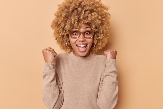 Prise de vue en intérieur de gestes de femme joyeuse émotionnelle excitée avec les poings s'exclame fort d'étonnement étant plein de joie regarde le match de football prend en charge le jeu préféré porte un pull décontracté isolé sur beige