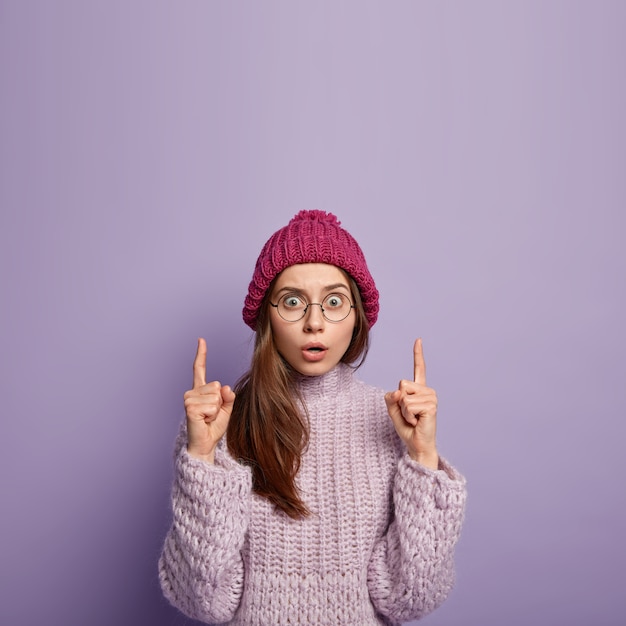 Prise de vue à l'intérieur d'une jeune femme caucasienne effrayée surprise au-dessus avec les deux index, porte des lunettes transparentes, des vêtements d'hiver, isolés contre le mur violet, de l'espace libre vers le haut