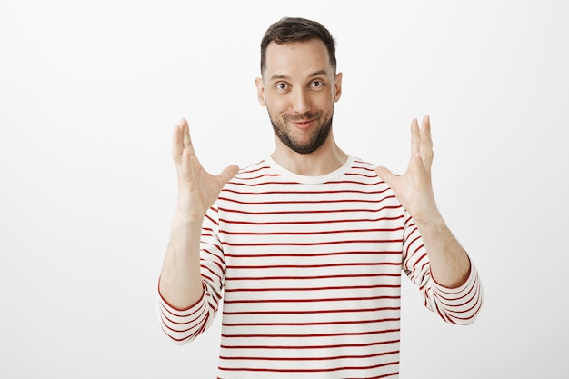 Prise de vue en intérieur d'un modèle masculin attrayant et heureux impressionné avec des poils, façonnant quelque chose de grand, décrivant la taille de l'article et la position