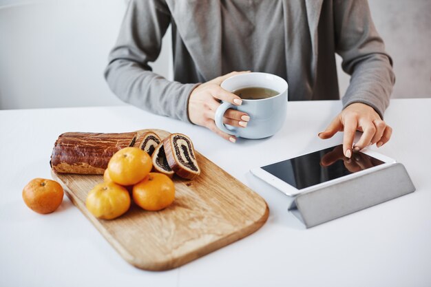 Prise de vue latérale des mains féminines avec manucure touchant la tablette numérique. Étudiante prenant son petit déjeuner avant d'aller à l'université, buvant une tasse de thé et mangeant des mandarines avec un gâteau roulé, elle se fait cuire